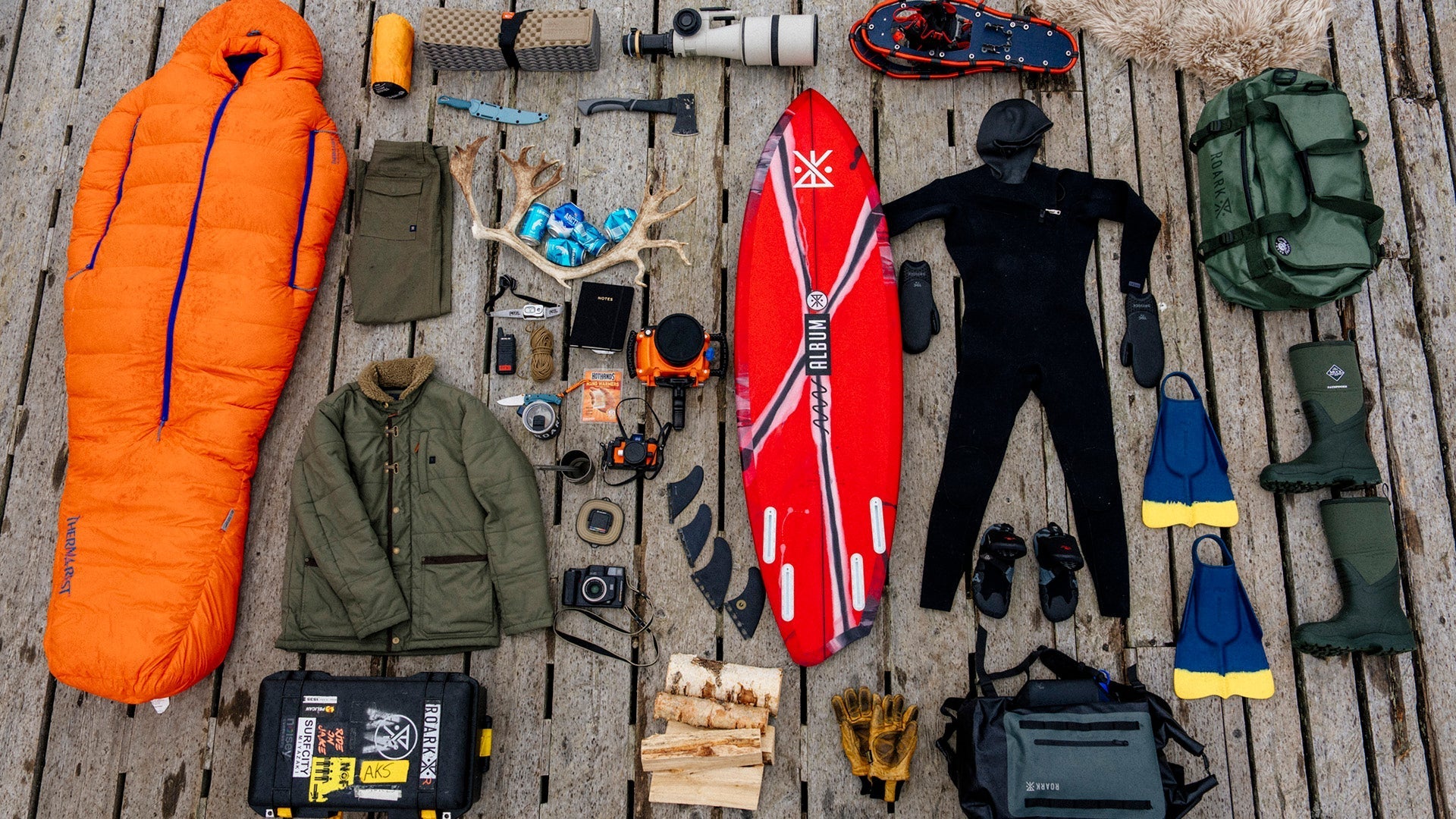 A flat-lay arrangement of essential winter surfing gear on a wooden dock. Includes a bright orange sleeping bag, wetsuit, surfboard, winter clothing, and various camping tools.